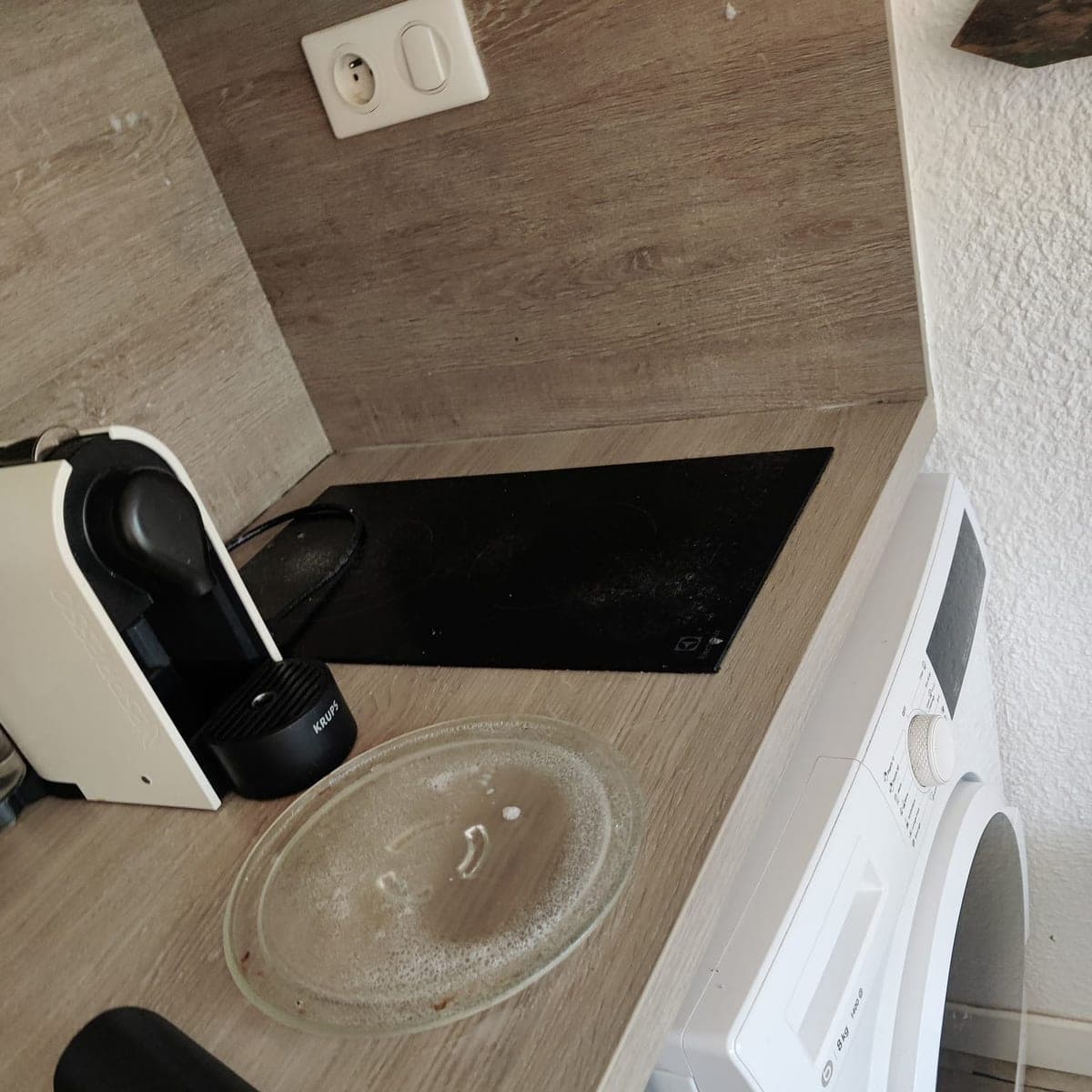 Plan de travail bois clair avec plaque de cuisson noire, machine à café et plateau de micro-ondes à l'état d'usage