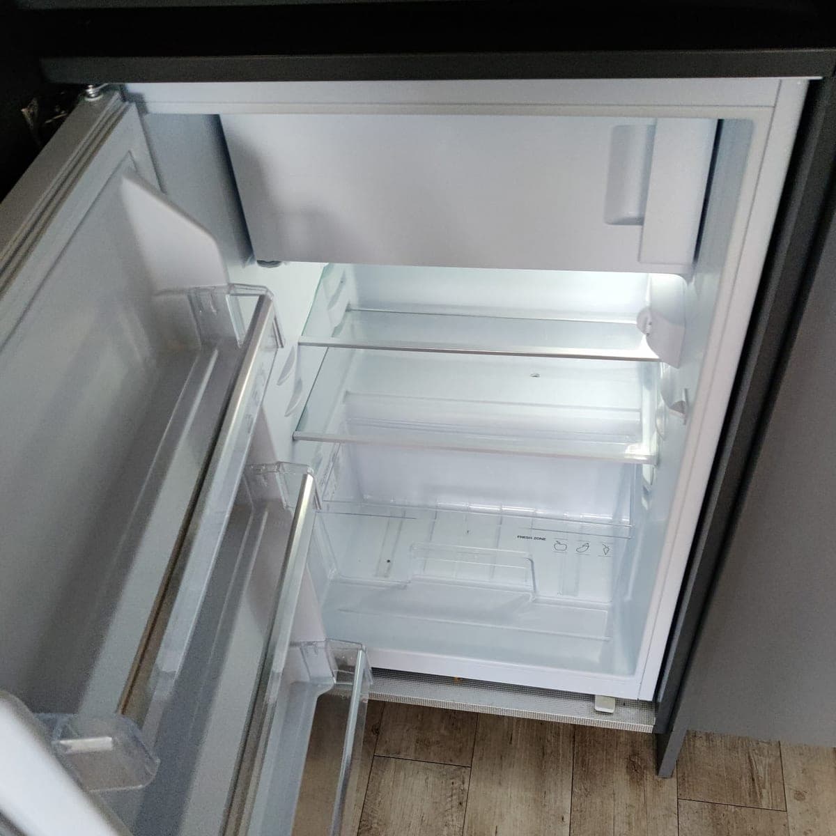 Réfrigérateur encastré ouvert, vidé, clayettes et bacs propres, sol imitation bois