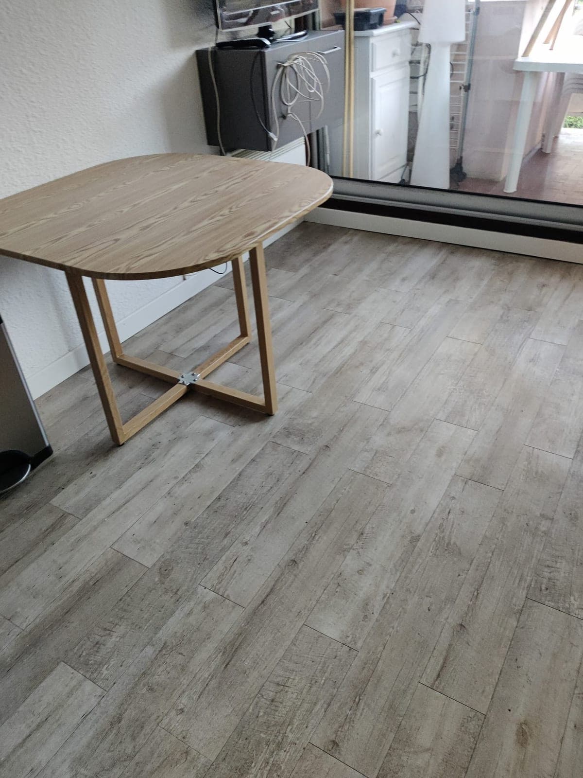 Table pliante en bois sur un sol imitation bois gris nettoyé, devant une baie vitrée