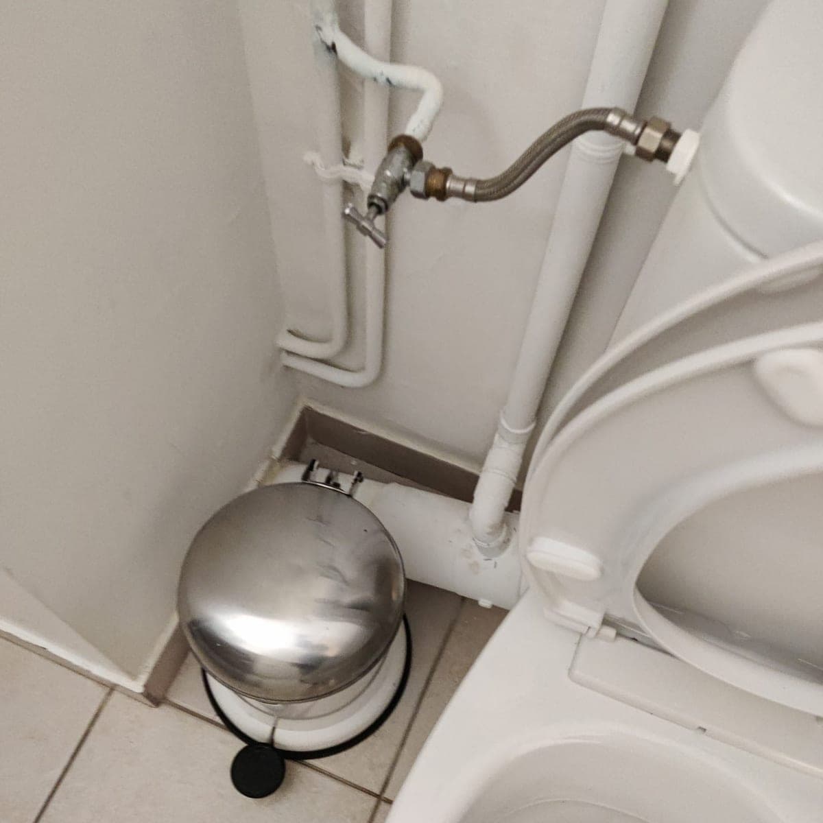 Coin derrière une cuvette de WC : tuyaux, plinthe et carrelage propres, avec une poubelle à pédale