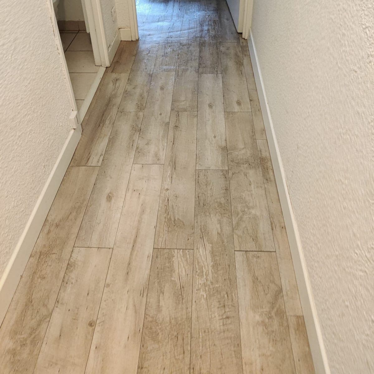 Couloir au sol imitation bois nettoyé, plinthes blanches, mur crépi, ouvert sur une pièce lumineuse