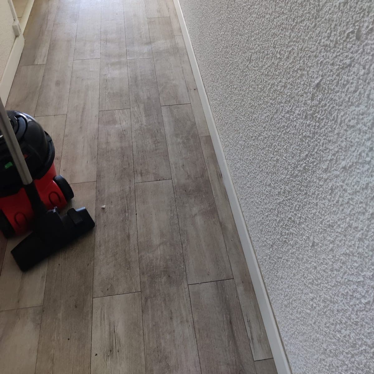 Couloir au sol imitation bois avec un aspirateur rouge en cours de passage, mur crépi blanc
