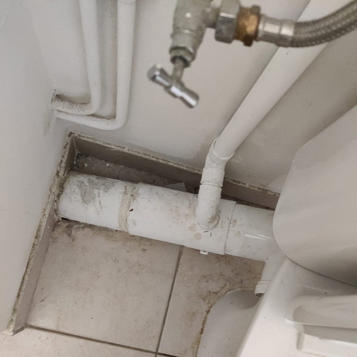 Coin derrière une cuvette de WC : tuyaux, plinthe et carrelage couverts de poussière et de traces grises