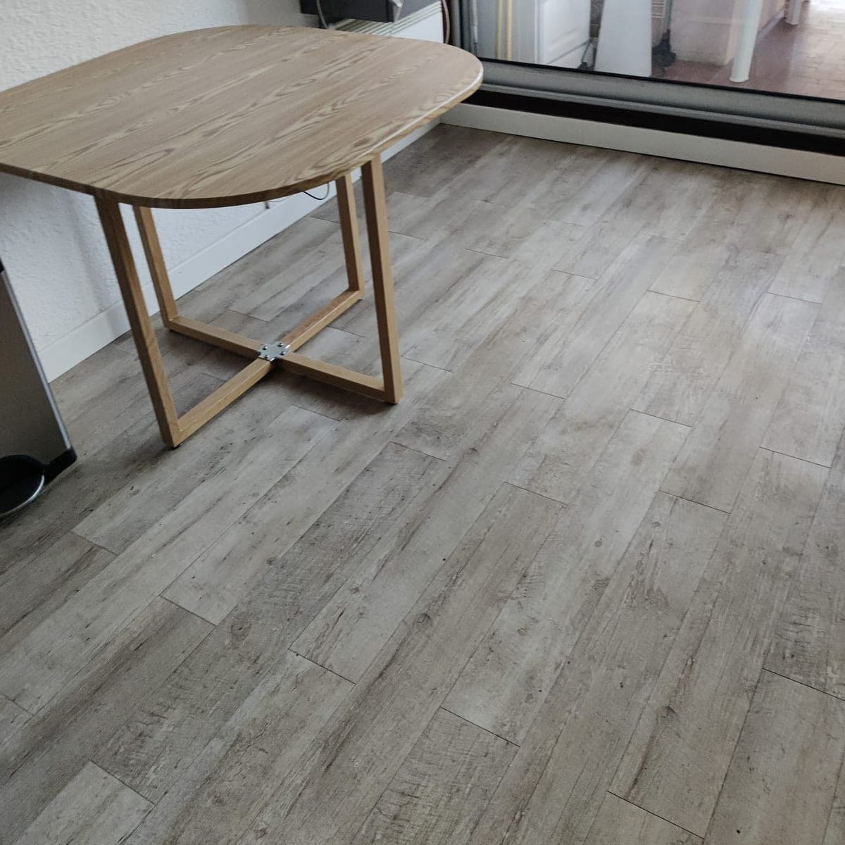 Table pliante en bois sur un sol imitation bois gris nettoyé, devant une baie vitrée